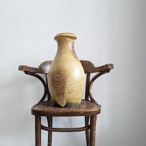 Puede incluir: Un jarrón de cerámica marrón y amarillo con una superficie texturizada se encuentra sobre una silla de madera con respaldo curvo.