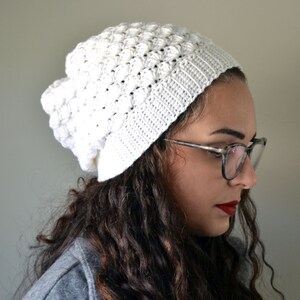 Pu&ograve; includere: Un berretto bianco all'uncinetto con un motivo a punto bobble testurizzato. Il berretto &egrave; indossato da una persona con capelli lunghi e ricci castani e occhiali.