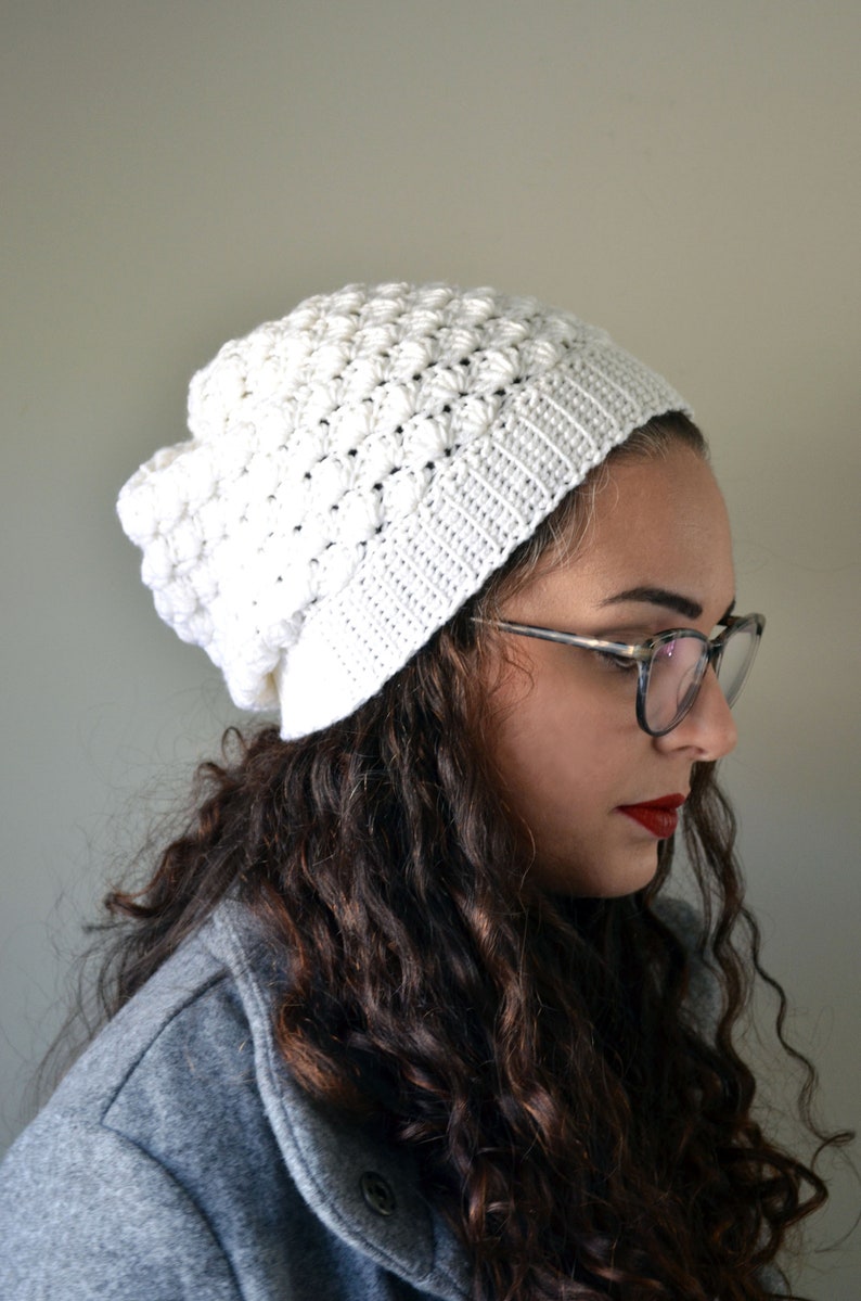 Pu&ograve; includere: Un berretto bianco all'uncinetto con un motivo a punto bobble testurizzato. Il berretto &egrave; indossato da una persona con capelli lunghi e ricci castani e occhiali.