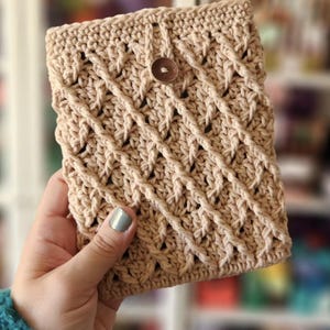 May include: A crocheted beige pouch with a brown button closure. The pouch is held in a hand with a light blue nail polish.