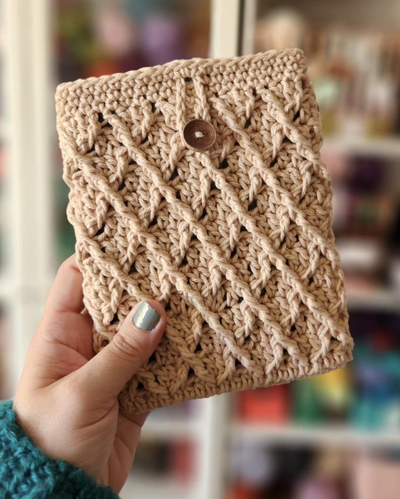 May include: A crocheted beige pouch with a brown button closure. The pouch is held in a hand with a light blue nail polish.
