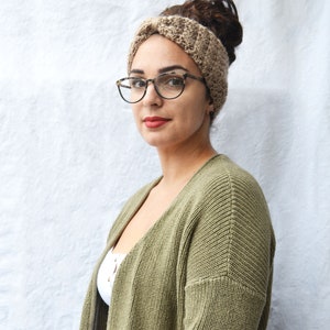 May include: A woman wearing a tan crocheted headband, tortoiseshell glasses, and a green cardigan. The headband is made of a textured yarn. The cardigan is a solid olive green color. The woman is looking directly at the camera.