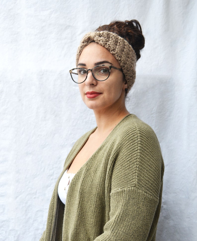May include: A woman wearing a tan crocheted headband, tortoiseshell glasses, and a green cardigan. The headband is made of a textured yarn. The cardigan is a solid olive green color. The woman is looking directly at the camera.
