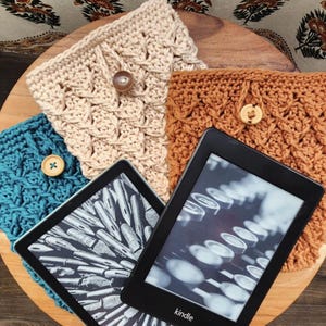 May include: Three crocheted Kindle covers in teal, beige, and brown with wooden buttons. The covers are displayed on a wooden surface with a Kindle e-reader displaying a black and white image of a typewriter.