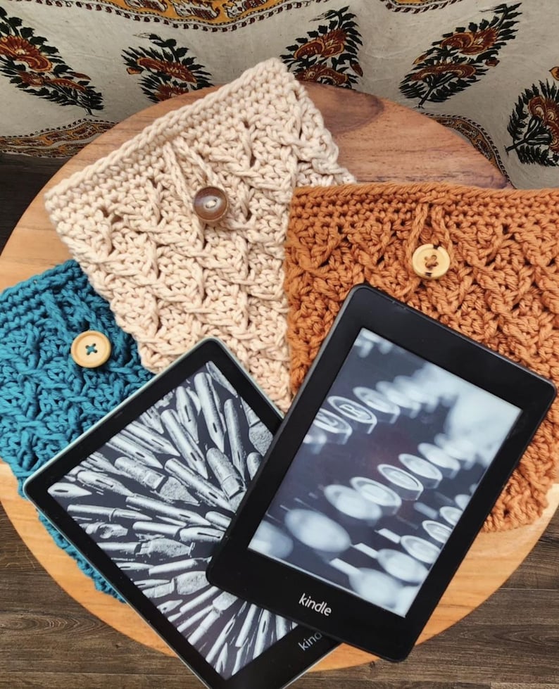 May include: Three crocheted Kindle covers in teal, beige, and brown with wooden buttons. The covers are displayed on a wooden surface with a Kindle e-reader displaying a black and white image of a typewriter.