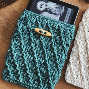 May include: A teal crocheted e-reader cover with a wooden button closure. The cover is made with a textured stitch pattern.