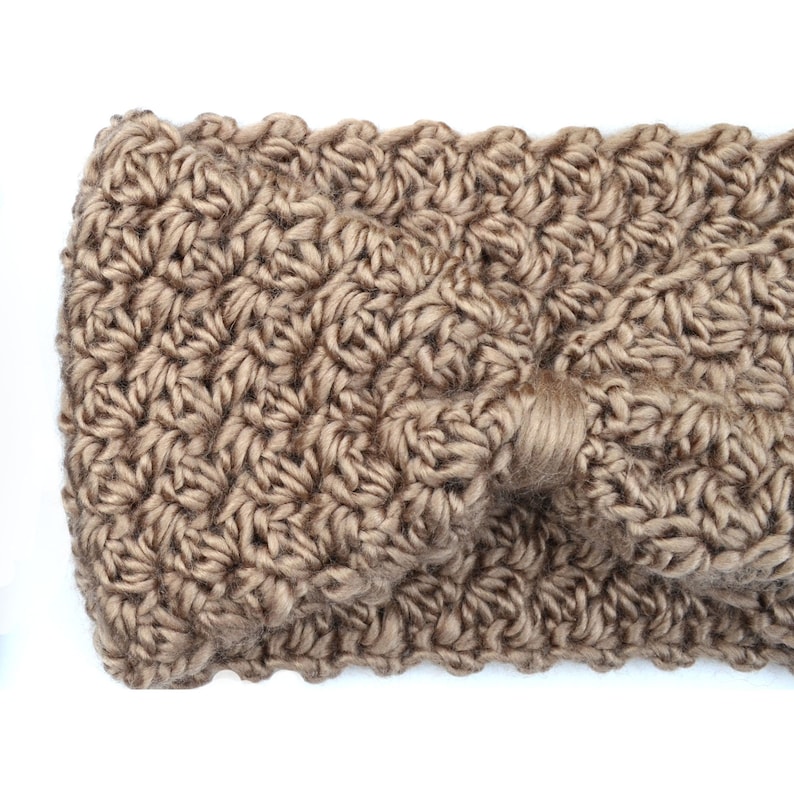May include: A close-up of a tan-colored, crocheted headband. The headband features a textured stitch pattern and a knotted detail on the front. The soft, warm accessory is likely made of wool or a similar yarn.