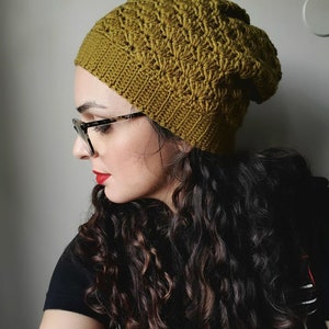 Pu&ograve; includere: Una donna con lunghi capelli castano scuro ricciuti indossa un berretto di lana verde lavorato a uncinetto. Indossa occhiali e ha il rossetto rosso.