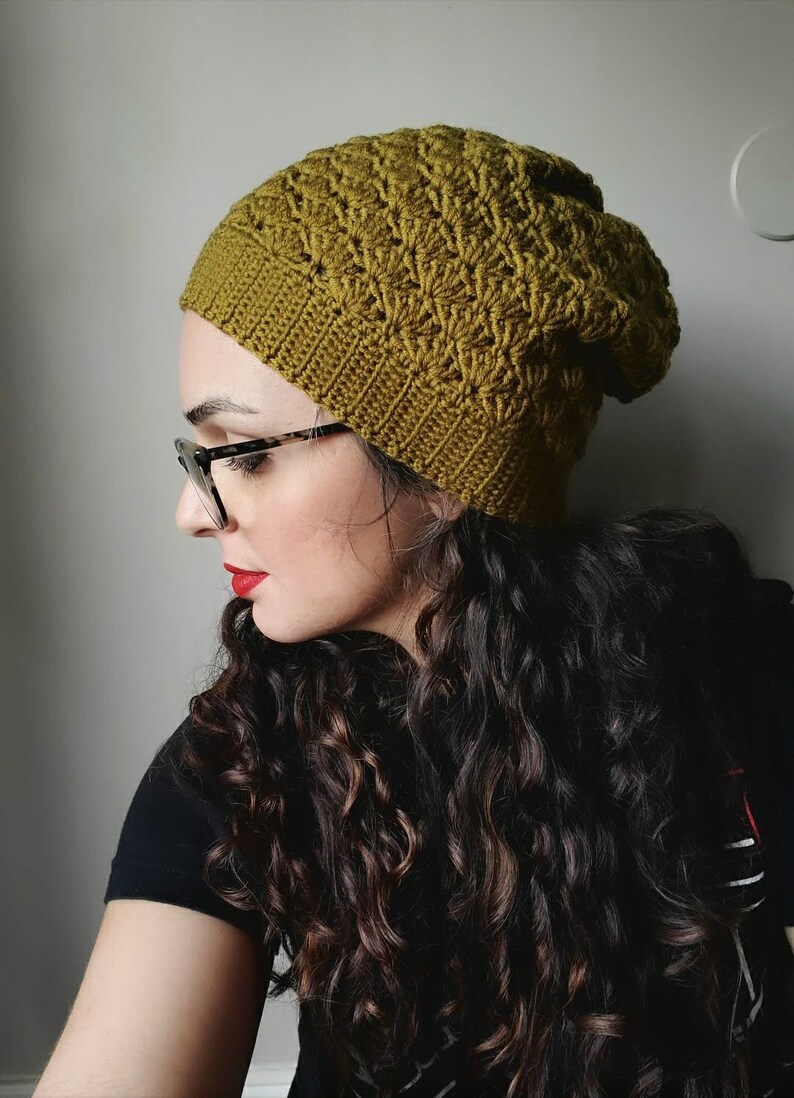 Pu&ograve; includere: Una donna con lunghi capelli castano scuro ricciuti indossa un berretto di lana verde lavorato a uncinetto. Indossa occhiali e ha il rossetto rosso.