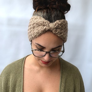 May include: A tan-colored, knitted headband with a bow detail is worn by a person. The headband is made of a textured yarn. The person is wearing glasses and a green cardigan. The background is a solid white.