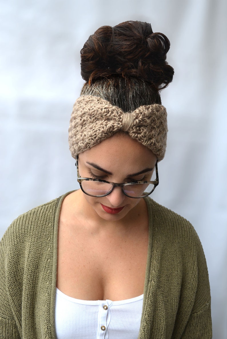 May include: A tan-colored, knitted headband with a bow detail is worn by a person. The headband is made of a textured yarn. The person is wearing glasses and a green cardigan. The background is a solid white.