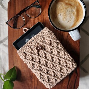 May include: A beige crocheted e-reader case with a wooden button closure. The case is on a wooden surface with a cup of coffee and glasses.