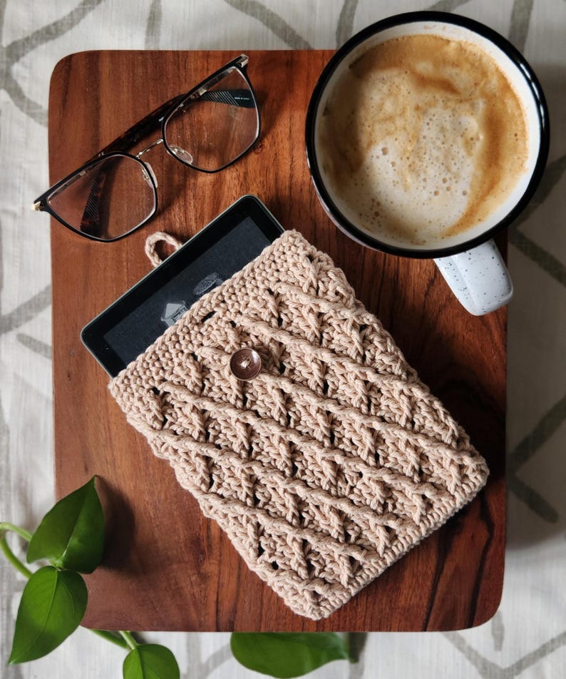 May include: A beige crocheted e-reader case with a wooden button closure. The case is on a wooden surface with a cup of coffee and glasses.