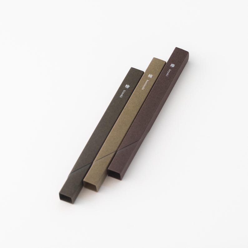 May include: Three brown, green, and dark brown incense sticks in their packaging. The packaging is made of paper and has Japanese characters printed on it. The sticks are arranged in a row, with the brown stick on top, the green stick in the middle, and the dark brown stick on the bottom.