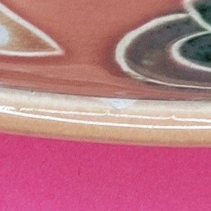 Może przedstawiać: Zbliżenie na dolną krawędź ceramicznej miski z brązową, pomarańczową, zieloną i białą glazurą. Miska ma lekko zaokrąglony brzeg.