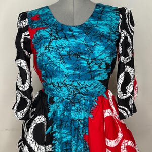 May include: A vibrant, patterned dress with a scoop neckline and long sleeves. The bodice features a turquoise and black design, while the sleeves and skirt incorporate black and white, and red patterns. The dress is displayed on a mannequin.