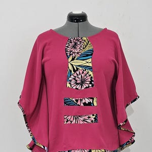 May include: A pink, short-sleeved top with a floral print panel on the front. The panel features a black background with pink, yellow, and blue flowers and leaves.