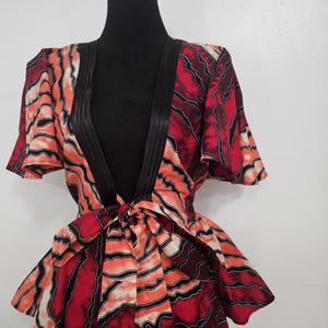 May include: A patterned wrap top in shades of red, orange, and black, displayed on a mannequin. The top features a V-neck, short sleeves, and a tie closure at the waist. The fabric has a wavy, abstract design.