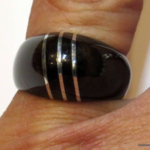 以下が含まれることがあります： 3本の銀色のストライプと2本の銅色のストライプが入った黒い指輪のクローズアップ。 指輪は滑らかで光沢があり、丸みを帯びた形をしています。 指輪は指にあります。
