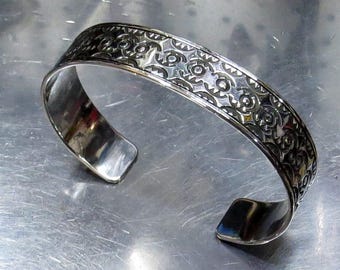 May include: A silver cuff bracelet with an intricate, repeating floral and geometric pattern. The open-ended bracelet is made of polished metal, reflecting light. The design is etched into the band, creating a textured surface. The bracelet is a piece of jewelry.