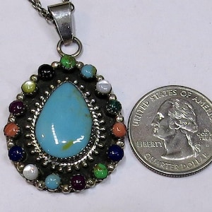 May include: A silver pendant necklace with a large, teardrop-shaped turquoise stone at the center, surrounded by a border of small, colorful stones. The necklace is displayed next to a U.S. quarter for size comparison.