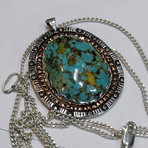 Puede incluir: Un collar con un colgante de plata y cobre con una gran piedra turquesa en el centro. El colgante tiene un diseño detallado alrededor de la piedra y está unido a una cadena de plata.
