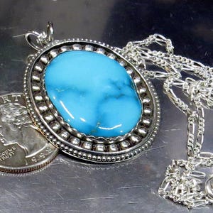 May include: Oval turquoise pendant necklace with a silver chain. The pendant features a large, bright blue turquoise stone set in a silver bezel with a beaded border. A U.S. quarter is next to the pendant for size comparison.