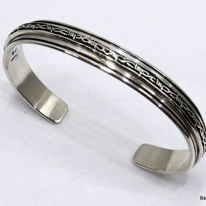 May include: A silver bracelet with a textured design of a repeating pattern. The bracelet is open and has a smooth, polished finish.