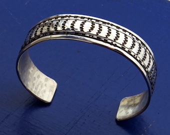 May include: A silver bracelet with a geometric pattern. The bracelet is made of hammered silver and has a polished finish.