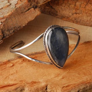 May include: A silver cuff bracelet with a large, teardrop-shaped, dark blue stone. The stone is set in a silver bezel with a twisted rope design. The bracelet is displayed on a wooden surface.