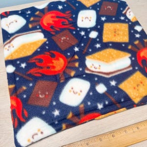 May include: A navy blue fleece blanket featuring a repeating pattern of s'mores, marshmallows, and flames. The s'mores have graham crackers, chocolate, and a toasted marshmallow. White stars are scattered across the blanket. A wooden ruler is in the foreground.