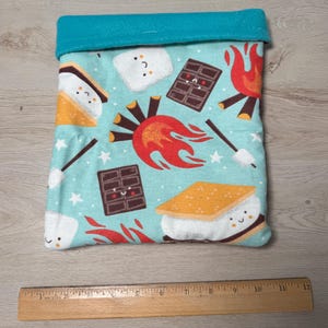 May include: A teal fabric pouch with a blue top edge. The pouch features a repeating pattern of cartoon s'mores, chocolate bars, campfires, and marshmallows. A wooden ruler is at the bottom of the image, measuring up to 12 inches.