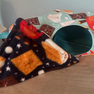 May include: A navy blue and orange s'mores-themed pet bed and matching triangular bed. The bed has a tunnel entrance and is decorated with marshmallows, chocolate bars, and a campfire design. The triangular bed has a s'mores design.