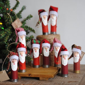 May include: A collection of 10 handmade Santa Claus ornaments made from repurposed shotgun shells. Each ornament is decorated with red, white, and green fabric, and has a white beard and a red hat. The ornaments are hanging from a brown wooden surface.