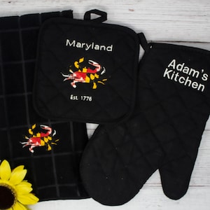 May include: Black kitchen towel and oven mitts with embroidered crabs and text. The towel has a grid pattern and the text "Maryland Est. 1776". The oven mitt has the text "Adam's Kitchen".