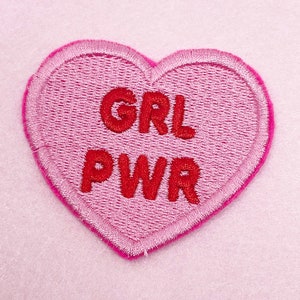 May include: Pink heart-shaped patch with red embroidered text that reads "GRL PWR".