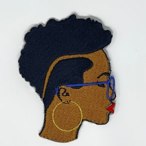 May include: Embroidered patch of a woman's face in profile. She has short, dark hair, brown skin, blue glasses, and large gold hoop earrings. Her lips are red.