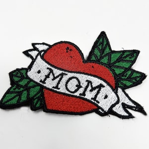May include: Red heart with a white banner that says "MOM" in a traditional tattoo style. The heart is surrounded by green leaves.