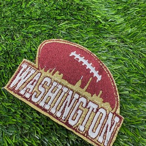 May include: A burgundy and gold embroidered patch featuring the word WASHINGTON in white block letters. The design includes a stylized football shape with white laces and a gold skyline silhouette integrated into the burgundy background.