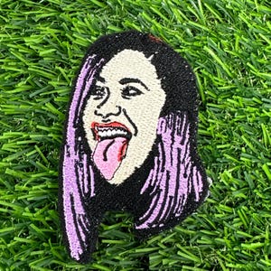 May include: Embroidered patch featuring a stylized portrait of a person with black hair and purple highlights. The person is sticking out their tongue and has fangs. The patch is on a green, grassy surface.