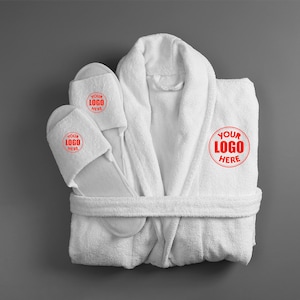May include: White terry cloth bathrobe and matching slippers set. The robe has a shawl collar and a waist tie. The slippers and robe feature a red circle with the text "YOUR LOGO HERE". The set is displayed on a gray background.