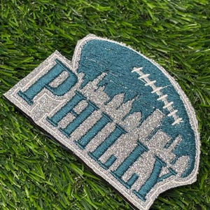 May include: A decorative embroidered patch featuring the word PHILLY in teal letters with a silver glitter outline. The design includes a silver glitter skyline silhouette and a white football lace graphic set against a dark teal background.