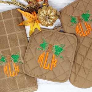May include: A brown kitchen set featuring a towel, pot holder, and oven mitt, all embroidered with an orange pumpkin design and the letters "NLK". The set has a quilted pattern and green leaf accents, perfect for fall.