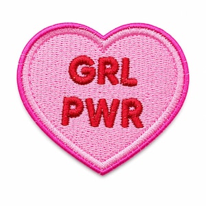 May include: A heart-shaped embroidered patch with a light pink background and a dark pink stitched border. The text GRL PWR is embroidered in bold red letters in the center of the heart, featuring a textured thread finish throughout the design.