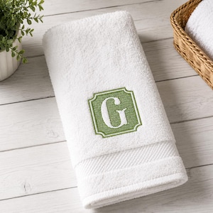 May include: A white cotton hand towel featuring a centered, embroidered green square patch with decorative scalloped edges. The patch contains a large, white, serif-style capital letter G. The towel has a textured border near the bottom edge.