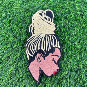 May include: Embroidered iron-on patch featuring a woman with a blonde bun hairstyle. The patch has a black outline and the woman's face is pink with red lips.