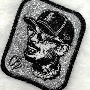 May include: A rectangular embroidered patch featuring a silver glitter background and a black stitched portrait of a person wearing a baseball cap and sunglasses. The design includes a signature in the bottom left corner and a black stitched border.
