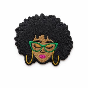 May include: An embroidered patch featuring a stylized face with a large black afro hairstyle, bright green cat-eye glasses, gold hoop earrings, and vibrant pink lips. The design is rendered in detailed thread work on a dark background.