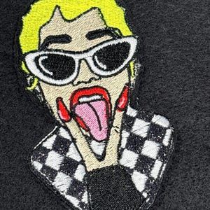 May include: Embroidered patch featuring a cartoon image of a person with yellow hair, white sunglasses, and red lipstick. The person is sticking out their tongue and making a peace sign. The patch has a black and white checkered pattern.