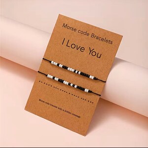 May include: Two Morse code bracelets with black and white beads are displayed on a brown card. The card reads "Morse code Bracelets" and "I Love You". The bottom of the card says "Morse code bracelet with a hidden message".